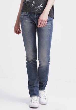 Person iført blå slim-fit jeans, en mørkegrå t-shirt med hvide guldsmedemotiver og hvide sneakers, stående foran en ensfarvet baggrund.