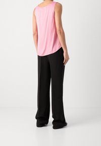 Woman wearing a sleeveless pink top and loose black trousers, standing with her back to the camera against a plain white background.