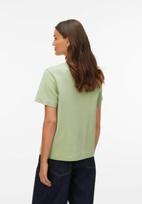 Jasnozielona koszulka z krótkim rękawem wykonana z miękkiej, gładkiej tkaniny. Luźny krój z prostym dołem i minimalistycznym wzorem, zestawiona z ciemnymi jeansami.