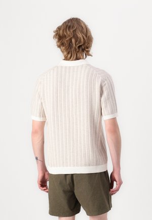 Person with wavy light brown hair wears a textured short-sleeve off-white sweater and olive green shorts, standing facing a plain background.