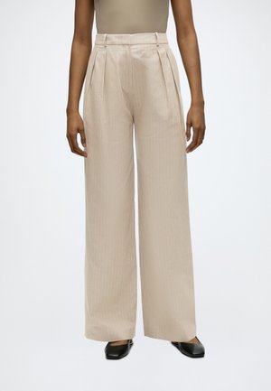 Beige bukser med høj talje og vide ben med tynde lodrette striber, sidelommer, bæltestropper, båret med sorte sko med firkantet tå og en tan ærmeløs top.