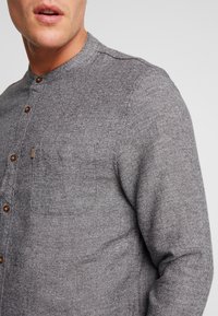 Hombre vistiendo una camisa de botones gris texturizada con collar mao y un bolsillo en el pecho con una pequeña etiqueta a rayas.