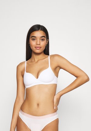 Soutien-gorge blanc rembourré avec fines bretelles, présentant une texture lisse et un décolleté plongeant. Associé à un slip de couleur claire affichant un bandeau avec un logo.
