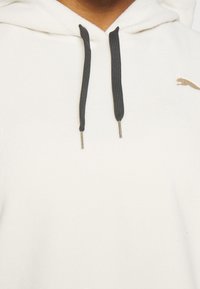 Gros plan d’un sweat à capuche blanc montrant des cordons noirs et un logo Puma doré partiel sur la partie droite de la poitrine.