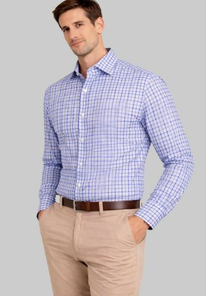 Homme portant une chemise à boutons à carreaux bleu clair et blanc, pantalon beige et ceinture marron, les mains dans les poches, sur un fond gris.