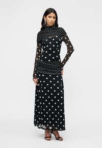 Kobieta ubrana w czarną prześwitującą sukienkę w grochy z długim rękawem, z dopasowanym szalem i czarnymi sandałami na wysokim obcasie, stojąca na jednolitym tle.