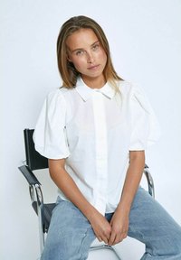 Blusa bianca con maniche a sbuffo e chiusura a bottoni, realizzata in un tessuto leggero. Indossata con jeans azzurri, seduta su una sedia in cromo.