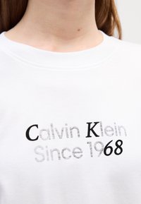 Witte katoenen t-shirt met een ronde hals. Bevat zwarte en grijze tekst: "Calvin Klein Sinds 1968" in verschillende lettertypen en groottes.