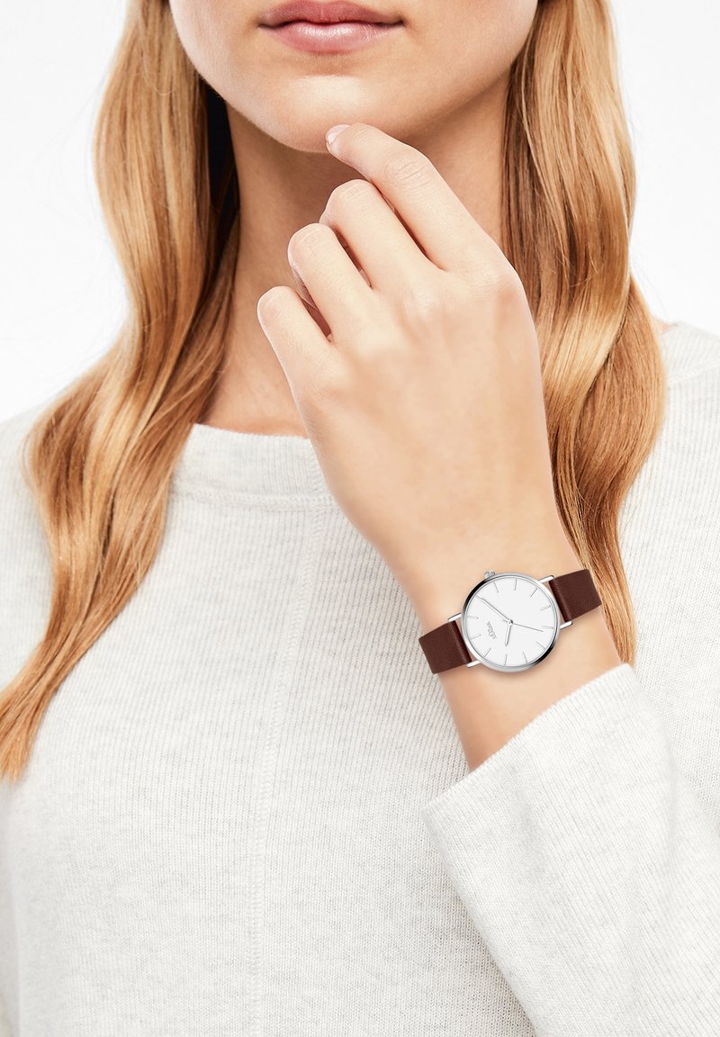 Femme aux longs cheveux châtain clair portant un pull gris clair et une montre-bracelet avec un cadran blanc et un bracelet en cuir marron.