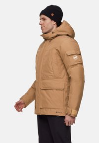 GORE-TEX isoleret jakke i tan, med hætte, flere frontlommer og justerbare manchetter. Glat tekstur og praktisk design.