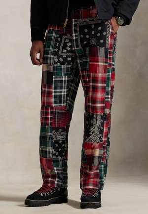 Mann trägt mehrfarbige Patchwork-Hose mit rot-, grün-, schwarz- und weißkarierten sowie Bandana-Mustern, schwarze Stiefel und eine schwarze Jacke.