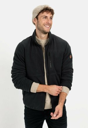 Schwarze Fleecejacke mit hohem Kragen, Reißverschluss-Brusttasche und strukturiertem beigefarbendem Pullover darunter. Model trägt eine hellfarbige Mütze.