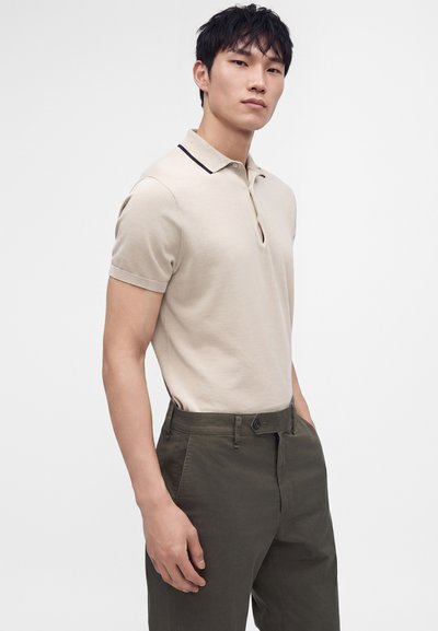 Jeune homme portant un polo beige à manches courtes avec une bande noire sur le col et un pantalon vert foncé, debout devant un fond blanc uni.