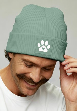 Man smiling and adjusting light green knit beanie with white paw print and heart logo on front, wearing white shirt against plain background.
