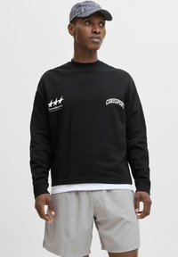 Hanorac negru cu logo alb "Coresport" și stele, purtat peste un tricou alb. Asortat cu pantaloni scurți gri deschis și o șapcă cu un model.