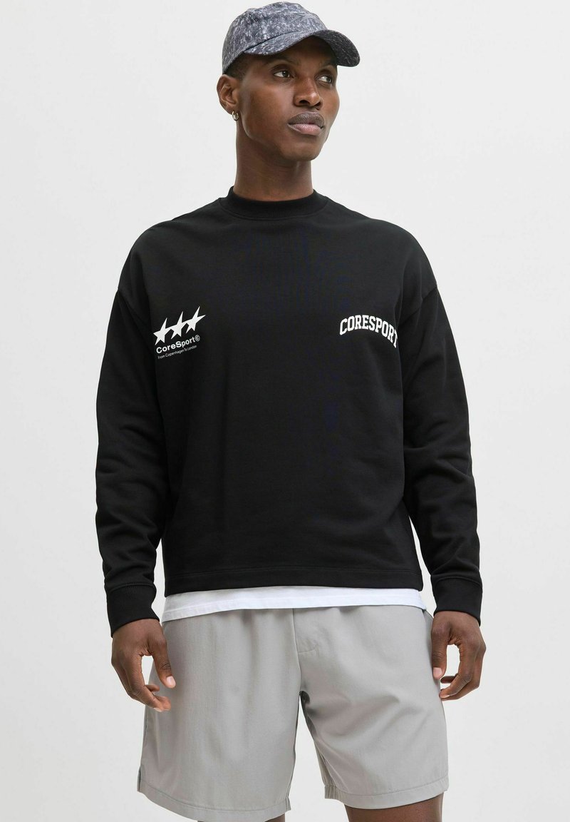 Hanorac negru cu logo alb "Coresport" și stele, purtat peste un tricou alb. Asortat cu pantaloni scurți gri deschis și o șapcă cu un model.