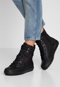 Bottes montantes noires en tissu et cuir, avec un design à lacets, une fermeture éclair latérale et une semelle en caoutchouc texturée. Jean partiellement visible.