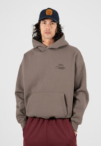 Haut à capuche marron en matériau doux, avec une poche avant et un logo brodé. Associé à un pantalon bordeaux et une casquette marine.