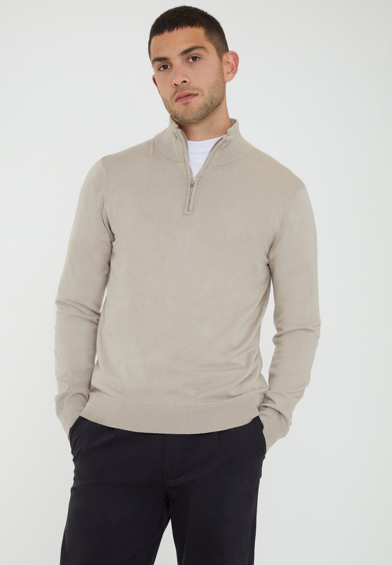 Beige knit pullover with a quarter-zip collar, long sleeves, and ribbed cuffs and hem, worn over a white shirt.