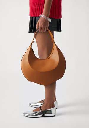 Brūnas ādas rokassoma ar izliekta formu un gludu tekstūru, kuru tur persona, kas ģērbjas sarkanā adītā džemperī un sudraba čībās.