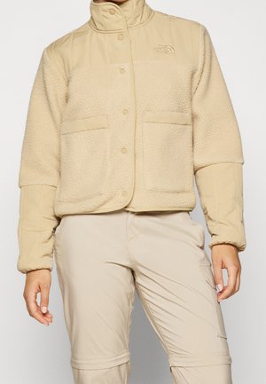 Beige fleecejakke med trykknapper og lappelommer båret sammen med matchende beige cargobukser på en stående person.
