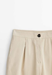 Pantaloni beige a vita alta con pieghe, tasche laterali, un bottone nero e zip frontale nascosta su uno sfondo bianco.