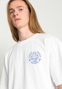 Hvit bomulls T-skjorte med et blått grafisk trykk av en frosk og teksten "EDWIN MUSIC CHANNEL" på fronten, kortermet, avslappet passform.