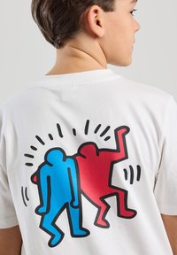 Valkoinen puuvillapaita, jossa on graafinen kuva kahdesta tyylitellystä hahmosta, yksi sininen ja yksi punainen, mustilla ääriviivoilla ja dynaamisilla viivoilla niiden ympärillä.