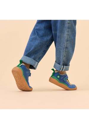 Kind trägt blaue und grüne Turnschuhe mit Gummisohlen, steht auf Zehenspitzen, trägt hochgekrempelte blaue Jeans vor einem beigen Hintergrund.