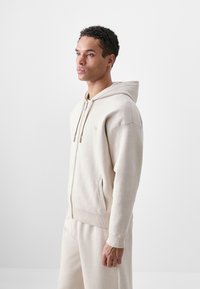 Bežinė zip-up palaidinė su priekinėmis kišenėmis, raukšlėtais rankogaliais ir su kapišonu, pagaminta iš minkštos medžiagos. Modelis stovi prieš paprastą foną.