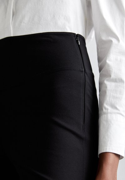 Black stretchy pants with a smooth texture, featuring a side zip fastening and flat seams. Paired with a white long-sleeve shirt.