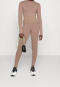 Brązowy prążkowany zestaw z długim rękawem i legginsy, dopasowany fason z okrągłym dekoltem. Model trzyma czarne i zielone taśmy do ćwiczeń.