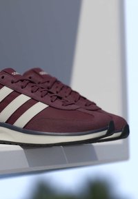 Paire de baskets Adidas bordeaux avec des bandes blanches exposée sur une étagère blanche sur un fond flou.