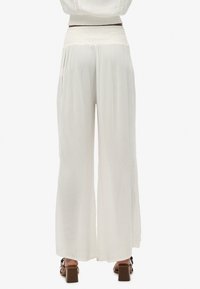 Pantalones blancos de pierna ancha hechos de una tela ligera con detalles texturizados y aberturas laterales, combinados con sandalias de tacón marrones.
