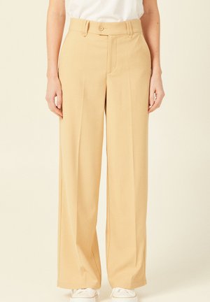 Beige weit geschnittene Hosen mit glatter Textur, hohem Bund, vorderen Falten und einem Knopfverschluss, kombiniert mit weißen Sneakern.