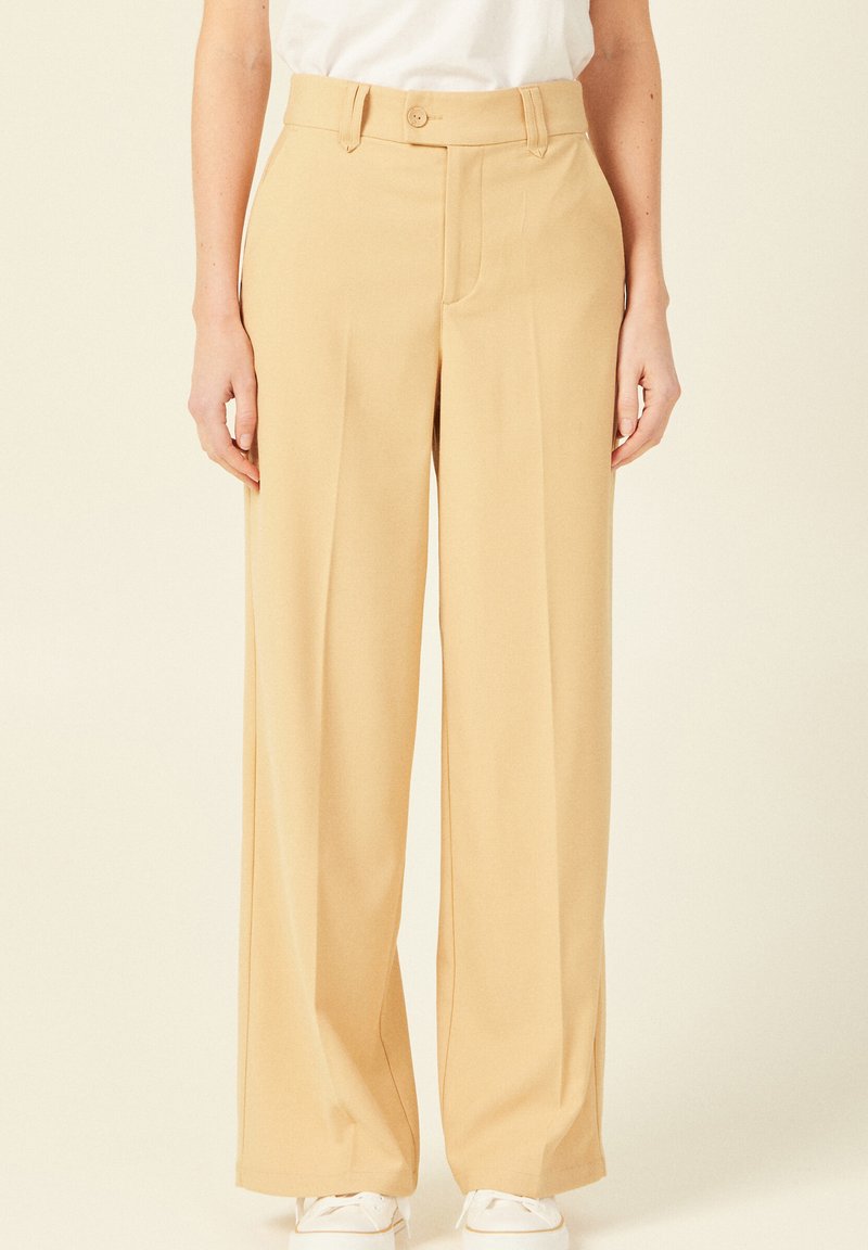 Beige wijde broek met een gladde textuur, hoge taille, plooien aan de voorkant en een knoopsluiting, gecombineerd met witte sneakers.