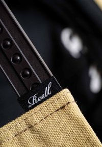 Khaki-Stofftasche mit schwarzem Etikett und weißem Text. Brauner Riemen mit kreisförmigen Akzenten. Nahaufnahme der Nähdetails und Textur.