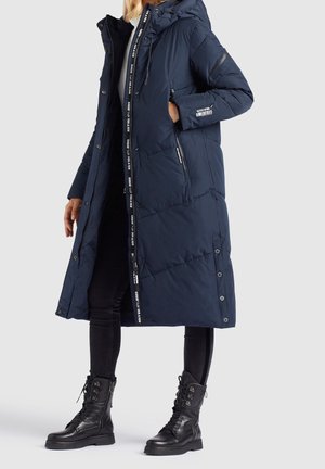 Femme portant un long manteau matelassé bleu marine avec capuche, des leggings noirs et des bottes de combat noires à lacets, debout devant un fond uni.