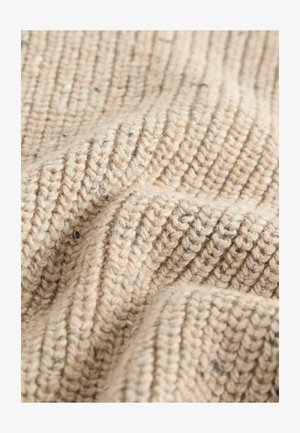 Beige kötött anyag, texturált, bordás mintával. A fonal sötétebb színű foltokat tartalmaz, amelyek vizuális érdekességet és mélységet adnak.