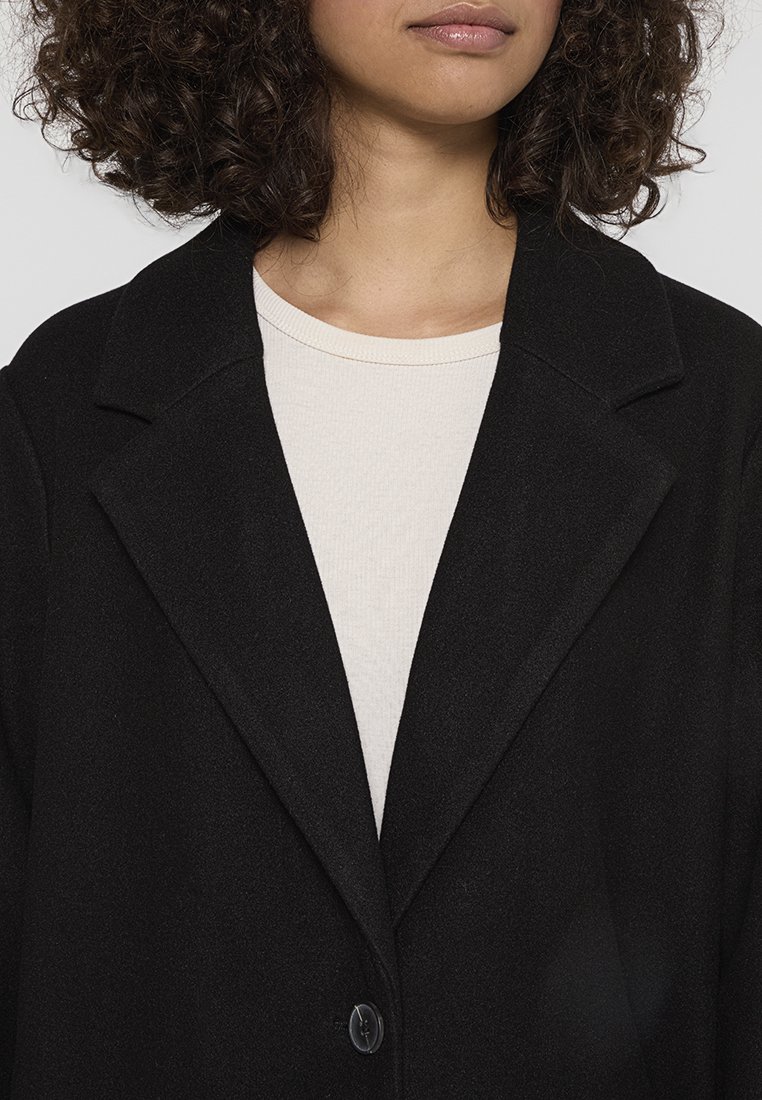 Zwarte blazer met een ingetogen kraag en enkele knoopsluiting, gedragen over een lichtbeige textuurtop, die de gladde uitstraling van de stof toont.