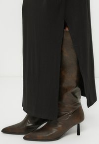 Καφέ μπότες με υφή μέχρι το γόνατο, με μυτερή μύτη και τακούνι στιλέτο, φορεμένες κάτω από ένα φαρδύ μαύρο ρούχο με πλαϊνή σχισμή.