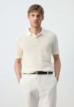 Man draagt een crèmekleurig gebreid poloshirt met korte mouwen, ingestopt in offwhite broek met een zwarte riem, staand tegen een effen lichte achtergrond.
