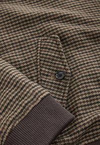 Rudas ir smėlio kolorijos houndstooth audinys su tamsiai ruda, raukšlėta apykakle ir juoda mygtuku prie priekinės kišenės. Minkšta tekstūra ir tvirta struktūra.