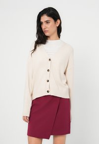 Μπεζ ζακέτα με λαιμόκοψη σε σχήμα V, πέντε σκούρα κουμπιά, ριμπ μανσέτες και άνετη γραμμή, σε συνδυασμό με μια μπορντό φούστα τύπου wrap. Μαλακή υφή, casual σχεδιασμός.
