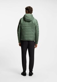 Uomo che indossa una giacca a vento verde trapuntata con cappuccio e pantaloni neri, in piedi di spalle contro uno sfondo luminoso e uniforme.