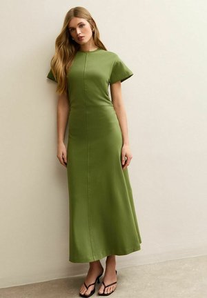 Vrouw met lang blond haar, gekleed in een strakke, olijfgroene maxi-jurk met korte mouwen en zwarte sandalen met hak, staand tegen een beige muur.