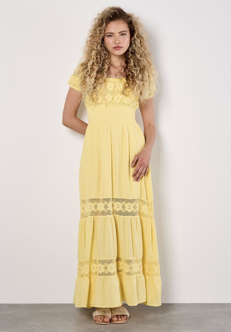 Jonge vrouw staat tegen een effen muur, gekleed in een gele maxi-jurk met kantdetails, met golvend haar en een neutrale uitdrukking.