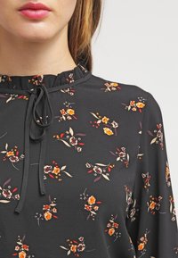 Svart blus med blommönster i orange, röd och krämfärgad. Har en rynkad krage och en snörning vid halsringningen, tillverkad av lättviktsmaterial.