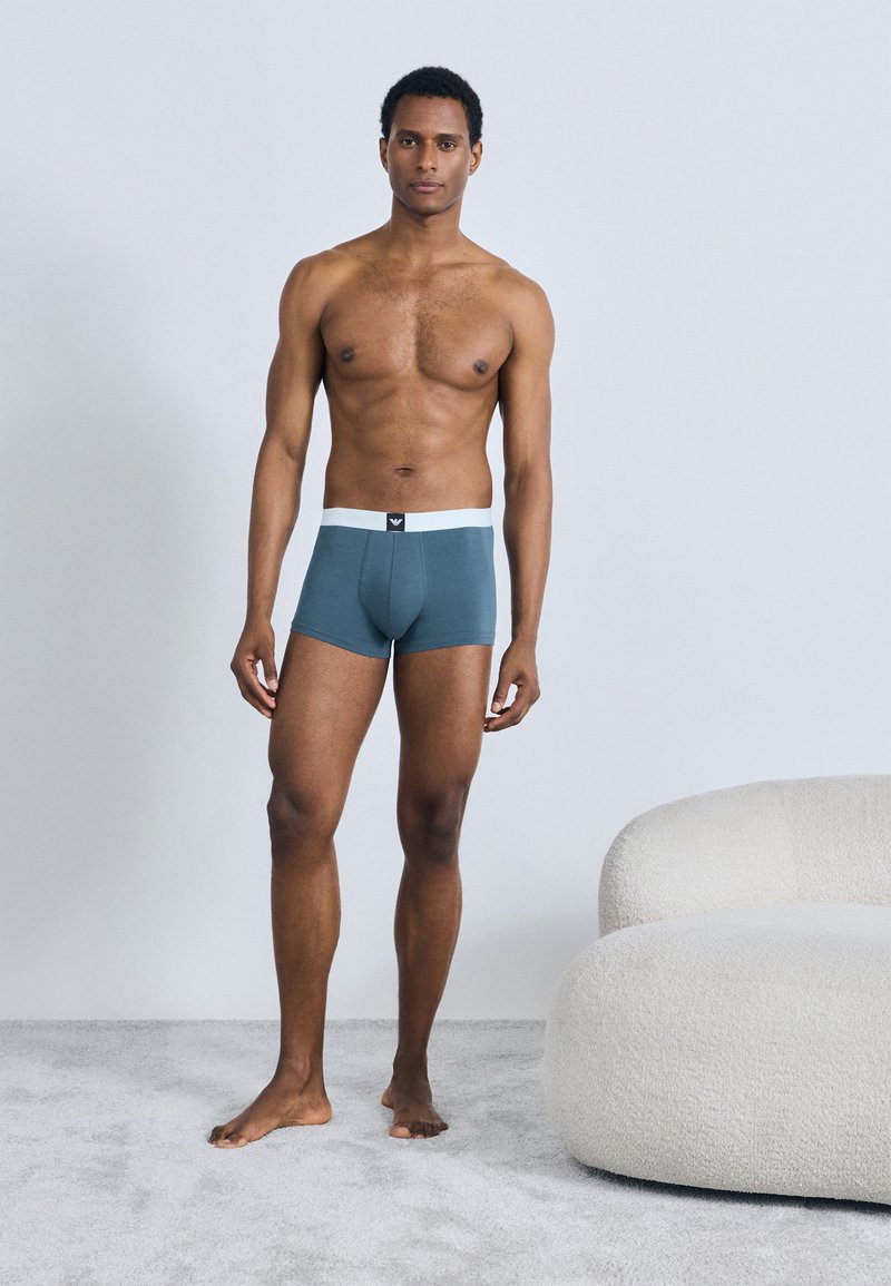 Vyrų bokserinės trumpikės žalsvai mėlynos spalvos su balta elastine juosta liemenėje. Audinys atrodo lygus ir glaudžiai prigludęs. Modelis stovi basas ant švelnios pilkos spalvos kilimo.