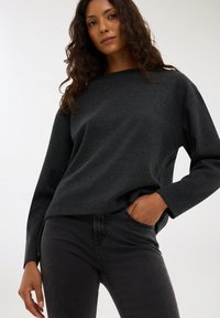 Ciemnoszary, miękki sweter o teksturowanej powierzchni z dekoltem typu łódka i długimi rękawami, zestawiony z dopasowanymi ciemnymi dżinsami. Minimalistyczny design, bez wzorów.